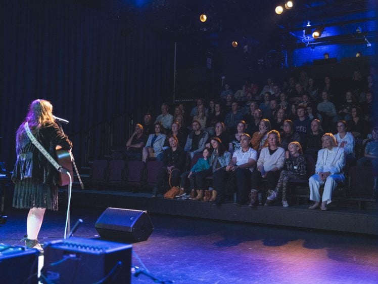 Image of A musician plays guitar and sings on stage under blue lights to an attentive, seated audience in a dark theatre. The audience appears engaged, watching the performance intently.