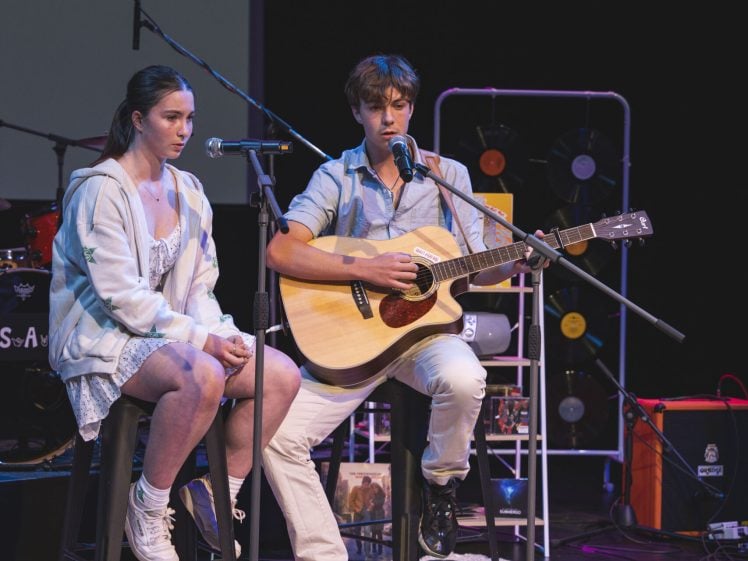 Image of A young man plays an acoustic guitar and sings into a microphone on stage, whilst a young woman sits beside him on a stool. Both appear focused, with musical equipment and stage lights visible in the background.
