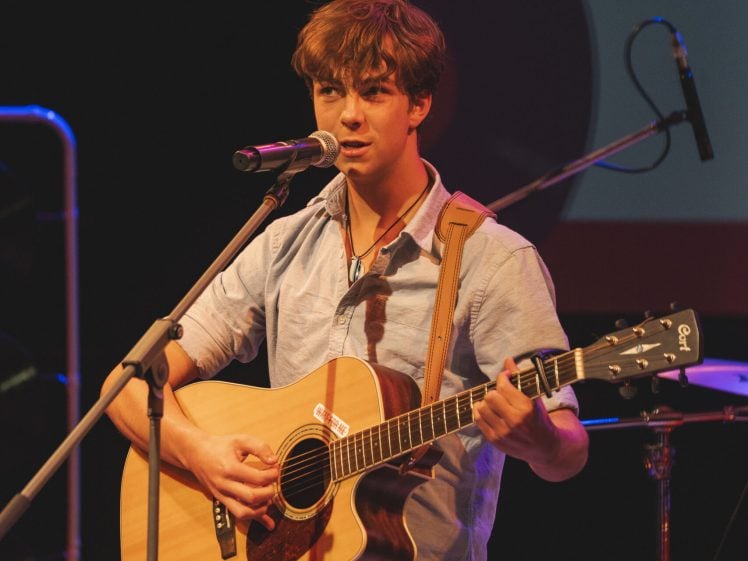 Image of A young man stands on stage playing an acoustic guitar and singing into a microphone. He wears a light blue shirt and beige trousers, and stage lights cast a warm glow on him. Musical equipment is visible in the background.
