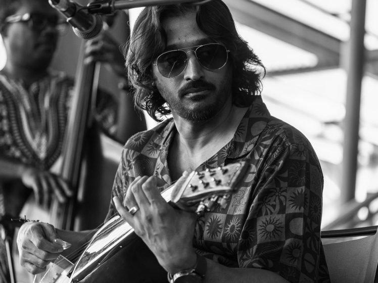Image of A man wearing sunglasses plays a stringed instrument, possibly a mandolin, whilst seated. Another musician in the background plays a double bass. The image is in black and white.
