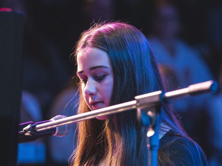 Image of A young woman with long brown hair plays the piano and sings into a microphone on stage, illuminated by colourful stage lights, with an audience blurred in the background.