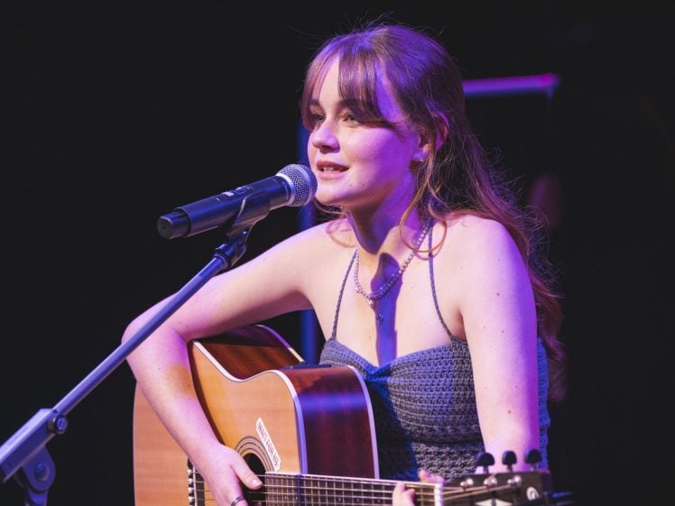 Image of A young woman with long brown hair plays an acoustic guitar and sings into a microphone on stage, wearing a blue top and white skirt, with stage lights illuminating her.