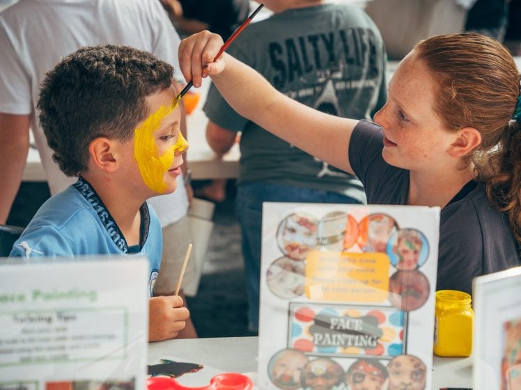 Image of A girl paints a boy’s face bright yellow at a face painting stall. They are smiling and surrounded by art supplies and signs describing the activity. Other children are visible in the background.