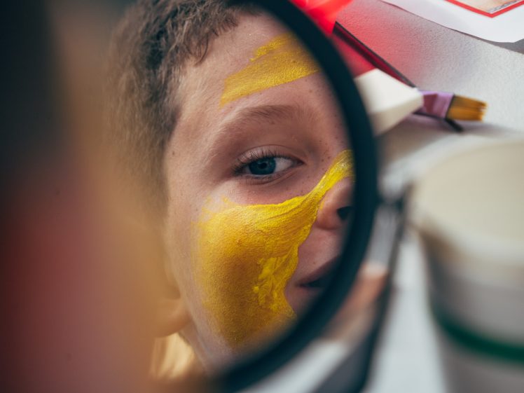 Image of A close-up of a child's face is seen in a round mirror, with yellow paint covering part of their cheek and forehead. Art supplies, including a brush and containers, are visible nearby.