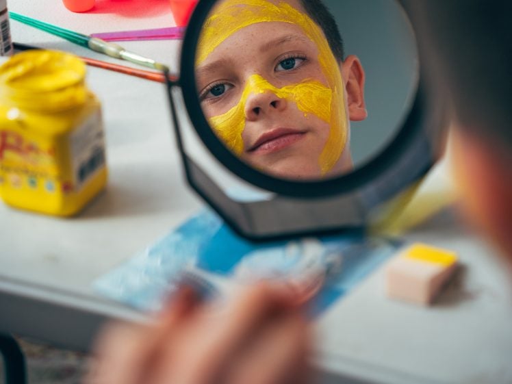 Image of A boy with yellow paint on his face looks at his reflection in a mirror. Art supplies and paint jars are scattered on the table in front of him.