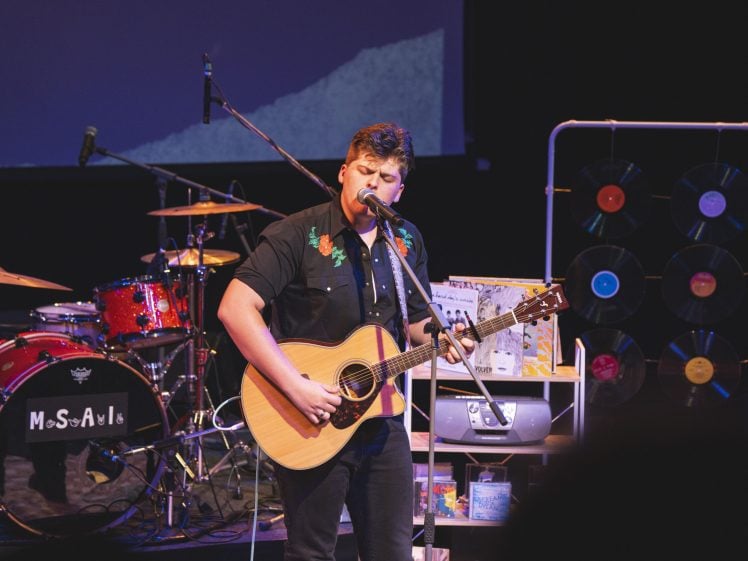 Image of A musician sings and plays an acoustic guitar on stage with a drum kit, vinyl records on a rack, and other musical equipment in the background under stage lighting.