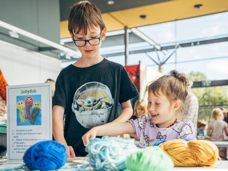 Image of A young boy and girl stand at a craft table with colourful yarn, smiling and engaged. A sign with instructions for making jellyfish crafts is visible. Other children and adults are in the bright, indoor background.