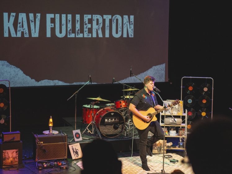 Image of A musician plays an acoustic guitar and sings on stage in front of a drum kit, amplifiers, and vinyl records, with KAV FULLERTON displayed on a large screen behind him.