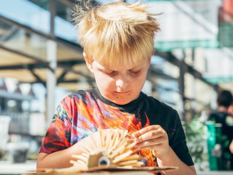 Image of A young boy with messy blond hair concentrates on crafting with wooden sticks at a sunlit table indoors. Bright sunlight streams through large windows behind him.