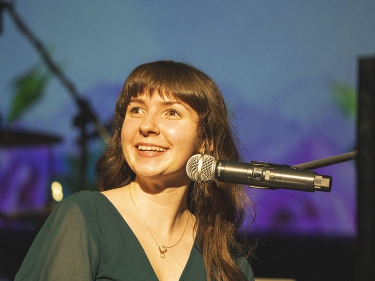 Image of A woman with long brown hair smiles while sitting at a keyboard and microphone on stage. She wears a green top and a gold necklace, with stage lights and a blurred background behind her.