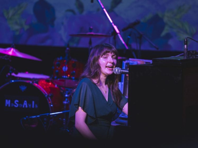 Image of A woman with long brown hair sings and plays the piano on stage under purple lighting, with a drum kit and microphone visible in the background.