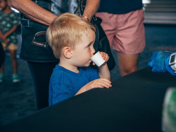 Image of A young blonde boy in a blue shirt drinks from a small paper cup whilst standing at a table. Adults stand nearby, and a person wearing blue gloves extends a hand towards him. The background is blurred.