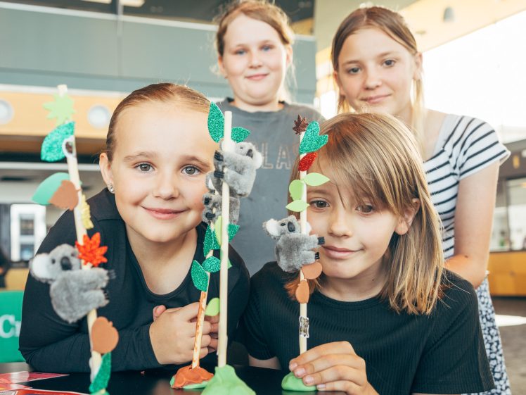 Image of Four children smile at a table displaying craft projects featuring toy koalas climbing homemade trees made from sticks, paper, and clay in a brightly lit indoor space.