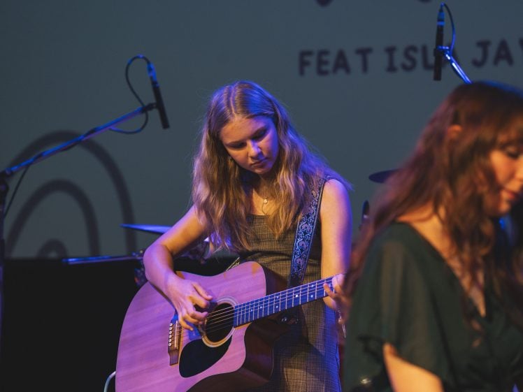 Image of A young woman plays an acoustic guitar on stage under stage lights, with another woman in the foreground and microphones and musical equipment visible in the background.