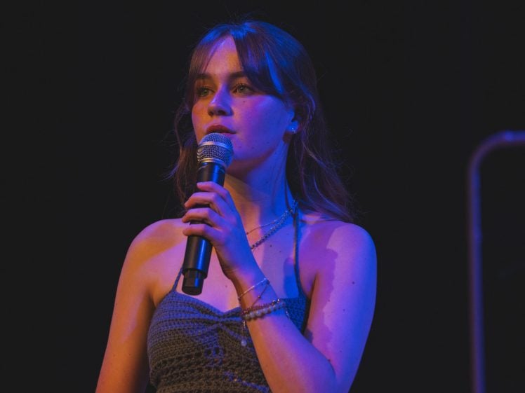 Image of A young woman stands on stage holding a microphone, illuminated by stage lights. She wears a sleeveless crochet top and a long skirt, looking thoughtful as she performs against a dark background.