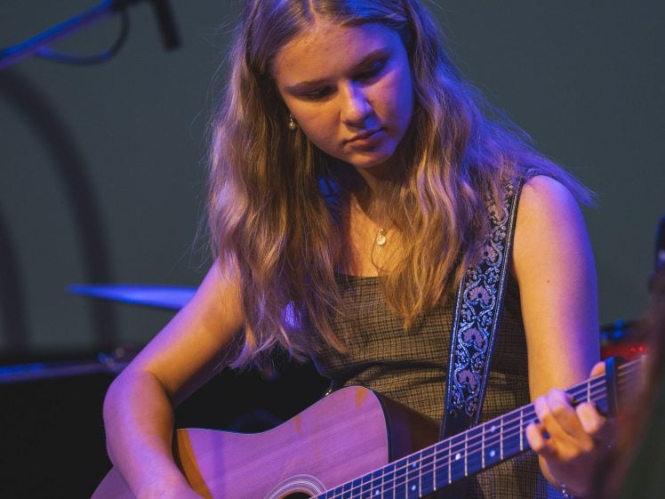 Image of A young woman with long, blonde hair plays an acoustic guitar on stage. She wears a sleeveless, checked dress and is focused on her performance under blue stage lighting. A microphone and drum kit are visible in the background.
