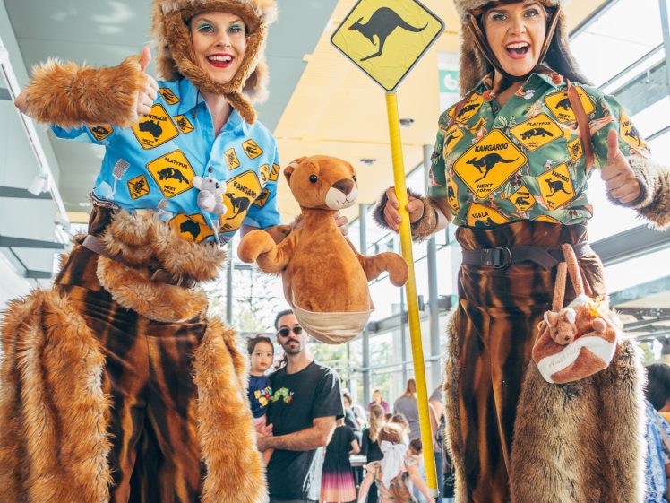 Image of Two people on stilts wear kangaroo costumes and blue shirts with Australian road signs, holding a kangaroo sign and a stuffed kangaroo. People and children watch in the background at an indoor event.