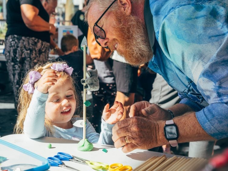 Image of A bearded man helps a young girl with a craft project at a table outdoors. The girl, smiling with a purple bow in her hair, looks excited as they work together, surrounded by art materials and other people.