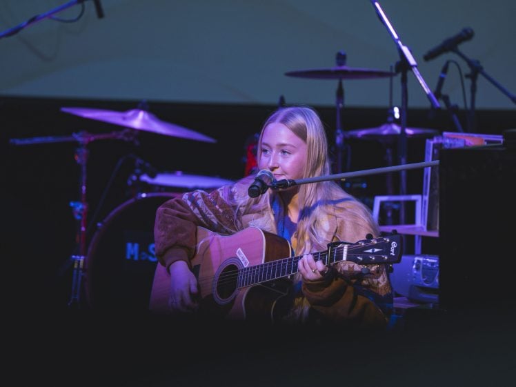 Image of A person with long blonde hair plays an acoustic guitar and sings into a microphone on stage, with drums and other musical equipment in the background. Purple stage lighting illuminates the scene.