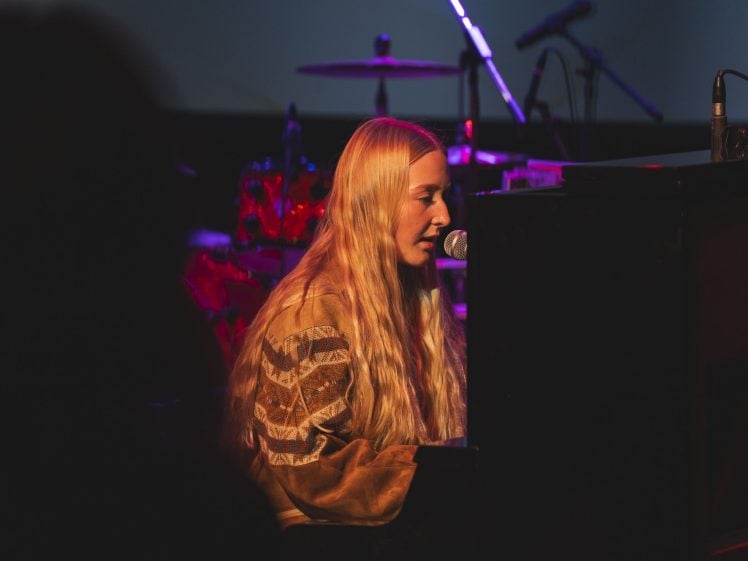 Image of A woman with long blonde hair plays the piano and sings into a microphone on a dimly lit stage, with drums and other instruments visible in the background.