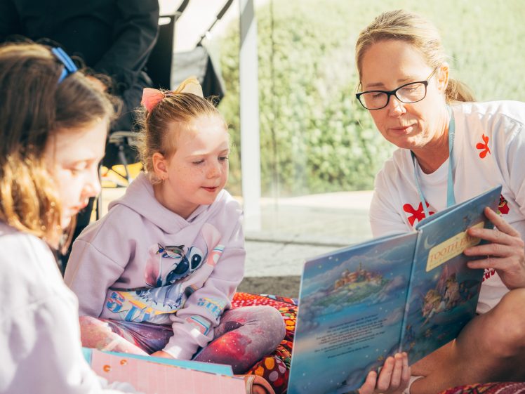 Image of A woman is reading a picture book to two young girls sitting beside her. They are indoors near a window, with sunlight coming in. The children listen attentively, and everyone looks engaged.