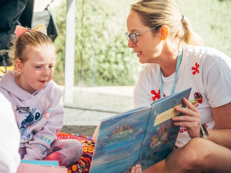 Image of A woman wearing glasses reads a children's book to a young girl with a pink bow in her hair. They are sitting together indoors, and the girl listens attentively. Sunlight comes through a nearby window.