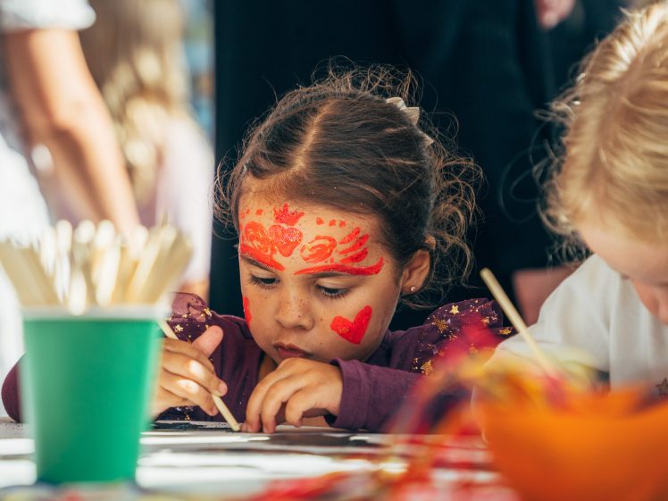 Image of A young child with red face paint concentrates on a craft activity at a table, surrounded by art supplies and another child. Bright colours and blurred background capture a lively, creative environment.