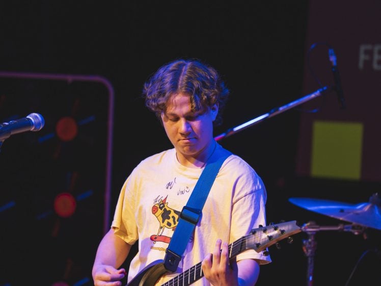 Image of A person with wavy hair wearing a graphic T-shirt plays an electric guitar on stage, with a microphone and drum kit visible in the background.