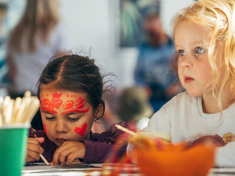 Image of Two young children sit at a table doing crafts. The child on the left has a red painted face and is focussed on drawing, whilst the blonde child on the right looks ahead thoughtfully. Art materials are visible on the table.