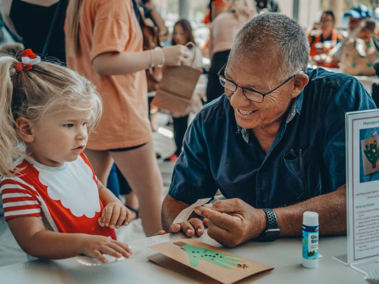 Image of An older man and a young girl work together on a craft project at a busy table. The girl wears a red and white outfit, and the man is smiling. Art supplies and a sign about making handprint Christmas cards are nearby.