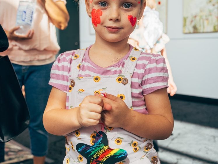 Image of A young girl with red face paint in the shape of a butterfly stands indoors, wearing a striped shirt and sunflower-patterned dungarees. An adult stands in the background holding a water bottle.