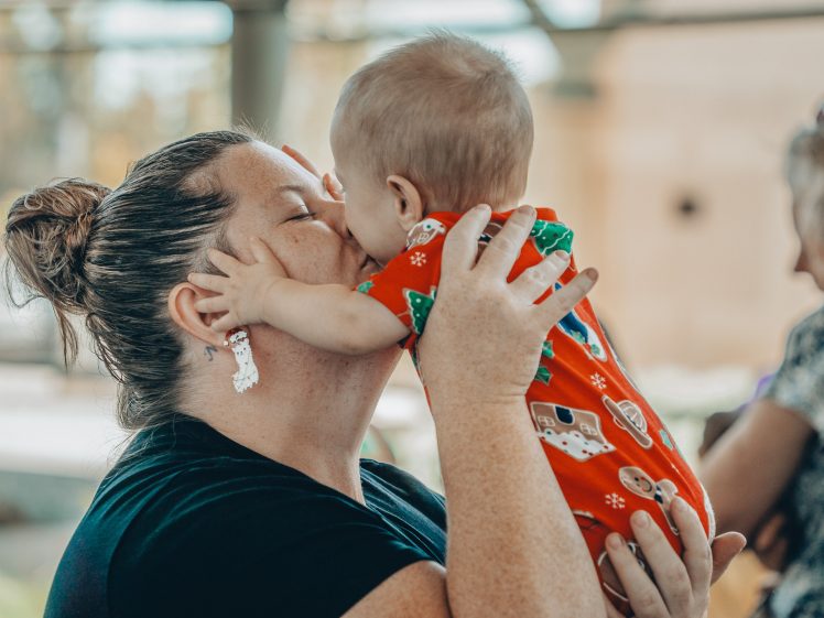 Image of A woman holds a baby close, smiling as the baby kisses her cheek. The baby wears a red outfit with holiday patterns. Another woman is visible in the blurred background.