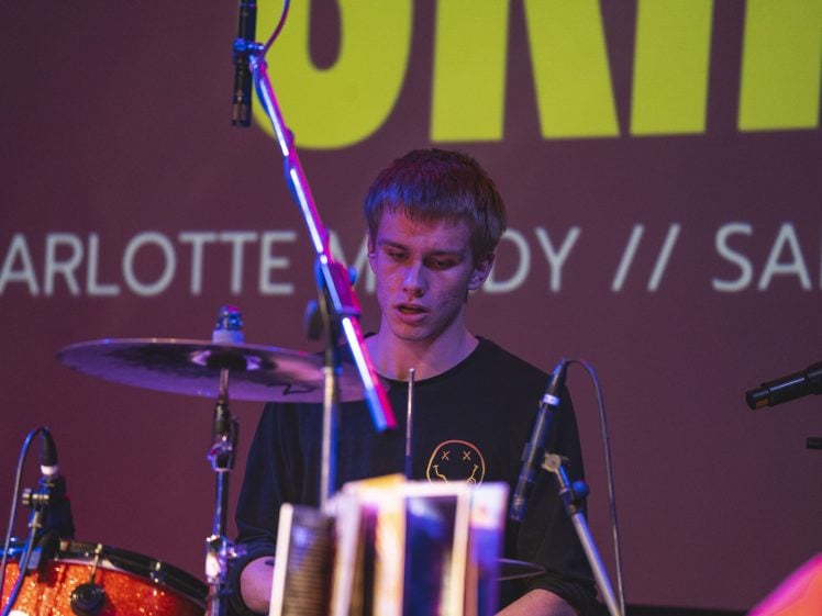 Image of A young drummer in a black shirt plays on stage, with a microphone and drum kit in front of him. A large, colourful backdrop with partially visible text is behind him.