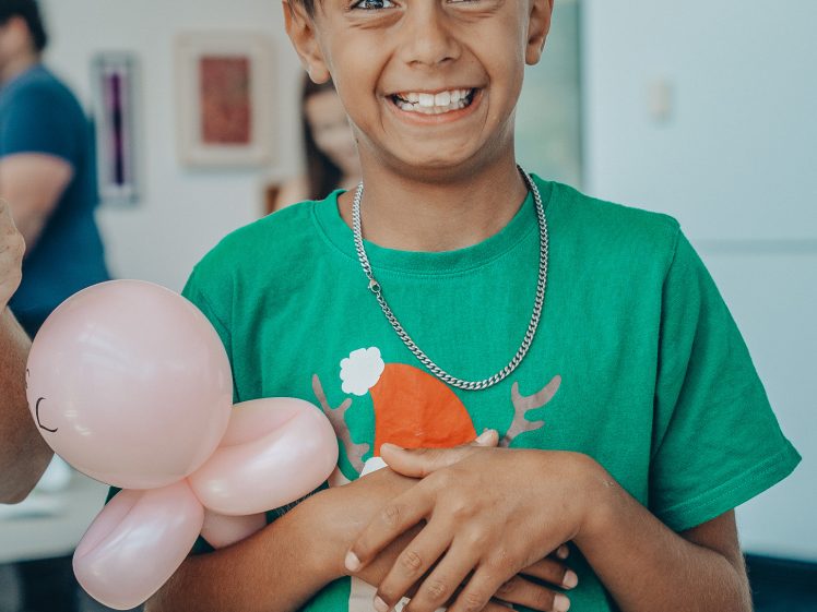 Image of A smiling boy with light brown hair, wearing a green Little Deer holiday shirt and a silver chain, holds a pink balloon animal indoors. Other people are visible in the background.