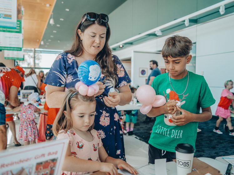 Image of A woman and two children with balloon animals participate in a craft activity at a busy indoor event. The little girl wears a balloon hat with a blue character, whilst the boy in green holds a balloon flower. People are visible in the background.