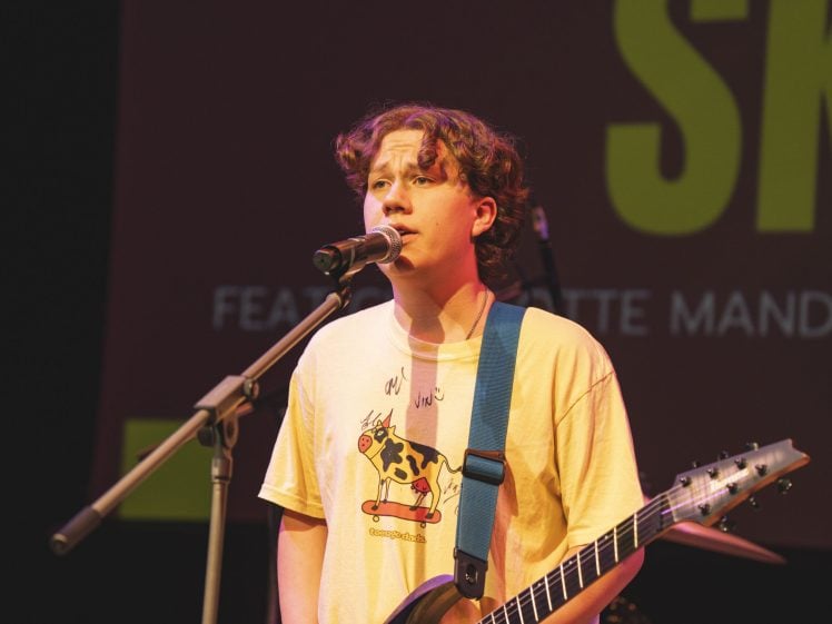 Image of A young person with curly hair sings into a microphone while playing an electric guitar on stage, wearing a yellow shirt with a cartoon cow graphic. Behind them, large yellow text is visible on a red screen.
