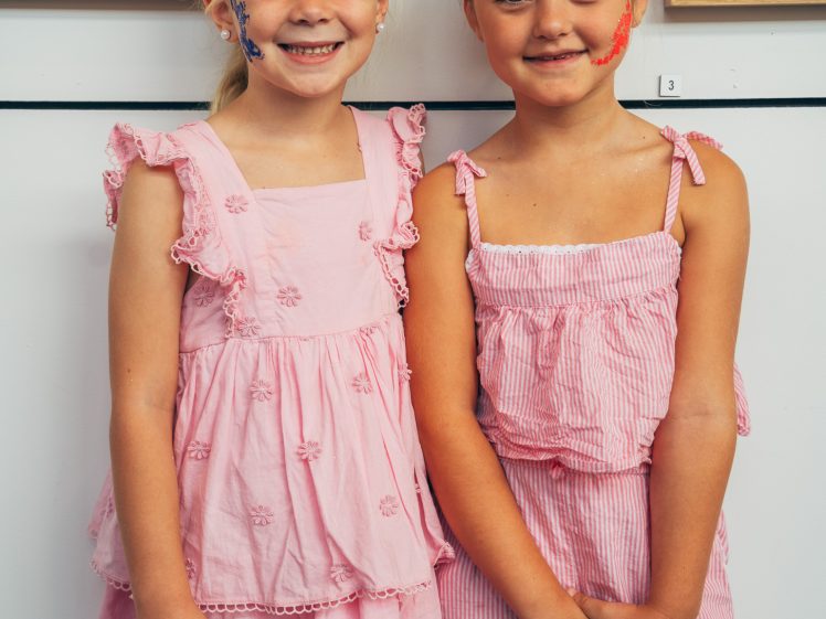 Image of Two smiling young girls with face paint stand side by side, both wearing pink outfits, in front of a white wall with colourful artwork hanging behind them.