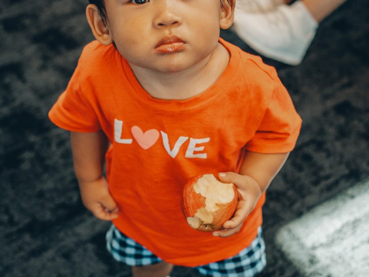 Image of A young child wearing an orange LOVE shirt and checked shorts looks up while holding a partially eaten apple, standing on a dark carpet with white trainers visible in the background.