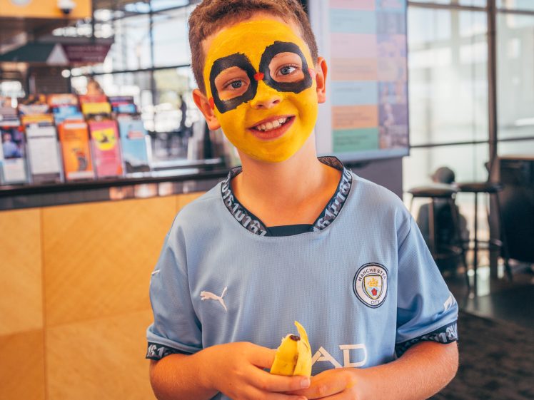 Image of A young boy wearing a Manchester City football shirt stands indoors, smiling and holding a peeled banana. His face is painted yellow and black, resembling a minion from the Despicable Me films.