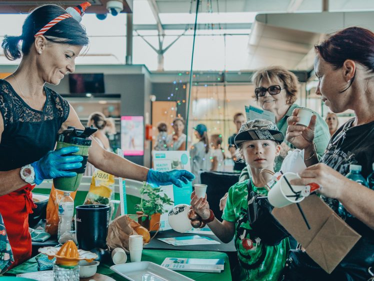 Image of A woman wearing gloves pours a green smoothie into cups at a busy indoor event. A child and adults holding cups and bags look on, surrounded by tables with informational materials and decorations.