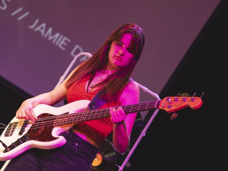 Image of A woman with long brown hair plays an electric bass guitar on stage under pink and purple lighting. She wears a red sleeveless top and black trousers. A screen with partially visible white text is in the background.