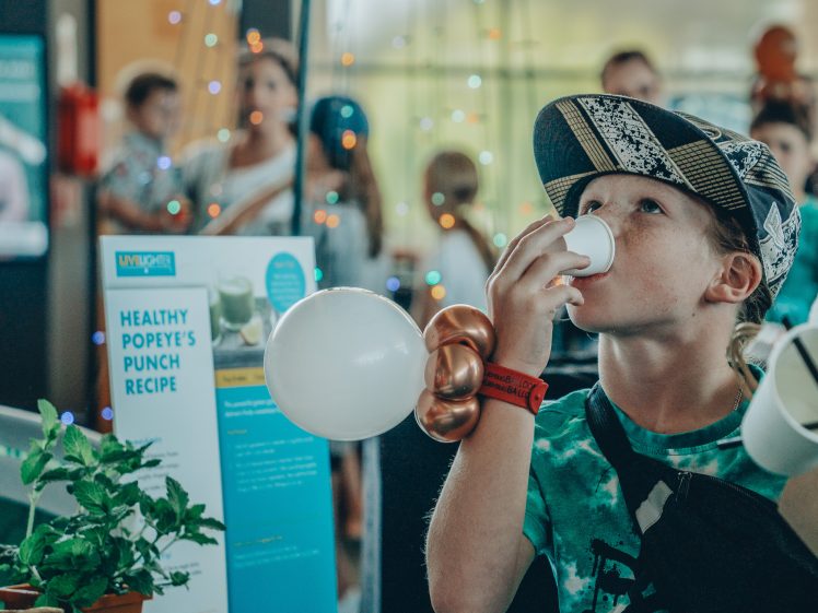 Image of A young child wearing a cap drinks from a small cup at an indoor event. A Healthy Popeye’s Punch Recipe sign, a mint plant, and balloon art are on the table. Several people are visible in the background.