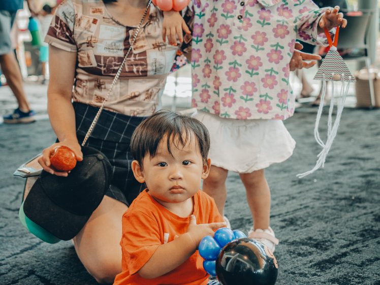 Image of A smiling woman kneels indoors with two young children. One child stands holding balloon animals and smiles, whilst the other sits on the floor holding balloon creations, looking at the camera.