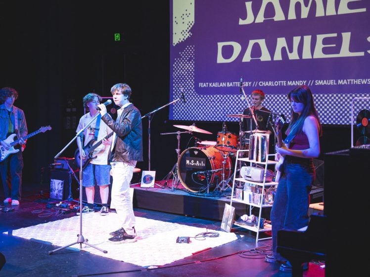 Image of A five-member band performs on stage with instruments; a screen behind them displays JAMIE DANIELS. The stage is lit with colourful lights, and the musicians play guitar, bass, drums, and sing.