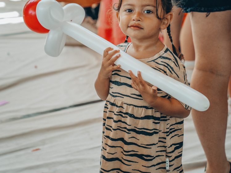 Image of A young girl in a striped dress holds a large balloon shaped like a rattle. She stands on a white surface with other people visible in the colourful, blurred background.