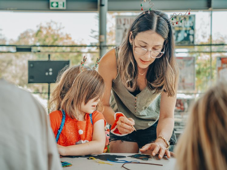 Image of A woman wearing glasses helps a young child with a craft project at a table. Both are wearing festive headbands. Other children sit nearby, and sunlight streams in through large windows in the background.