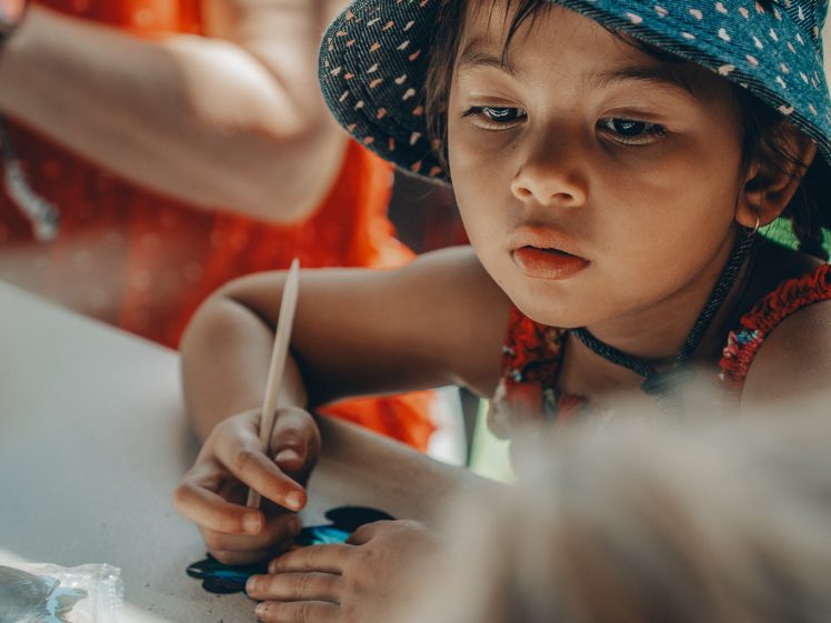 Image of A young child wearing a blue spotted hat focuses intently whilst painting at a table, holding a brush. An adult in a red shirt is seated nearby, partially visible in the background.