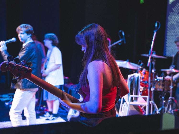 Image of A band performs on stage under colourful lights. A woman in a red top plays bass guitar in the foreground, while other musicians, including a singer with a microphone, stand in the background near drum and keyboard set-ups.