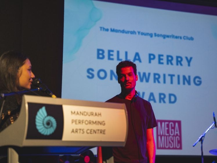 Image of A woman speaks at a lectern labelled Mandurah Performing Arts Centre while a man stands beside her. Behind them, a screen displays Bella Perry Songwriting Award and event sponsor logos.
