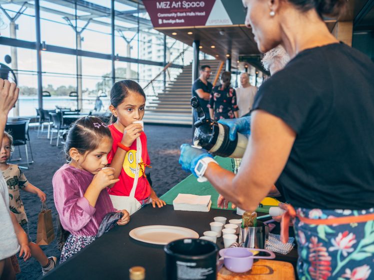 Image of Children and adults gather at a table, sampling drinks served by a woman wearing gloves in a spacious, sunlit indoor area with stairs and signs in the background.